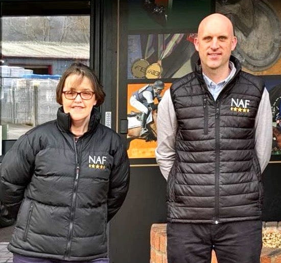 Two people wearing NAF branded jackets standing in front of a store with sports memorabilia.